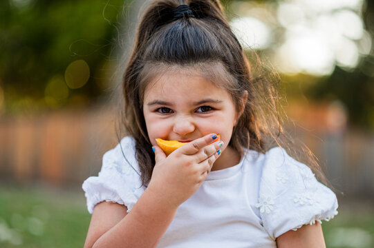 Young girl smiles while taking a bite of a mango slice in a garden during the afternoon