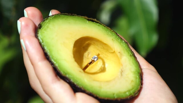 Woman's manicured hand holding a fresh, green half avocado containing a beautiful gold diamond engagement ring, a creative and unique marriage proposal concept with a healthy lifestyle twist