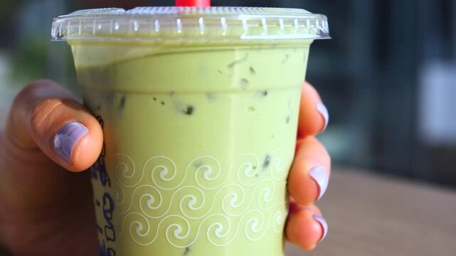 Close up of a woman's hand with purple nail polish holding a plastic cup of iced matcha green tea latte, slowly turning it to show the beverage with ice cubes inside a cafe