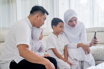 Obraz premium Muslim Family in White Traditional Attire Making a Video Call on Smartphone During Eid al-Fitr Celebration at Home