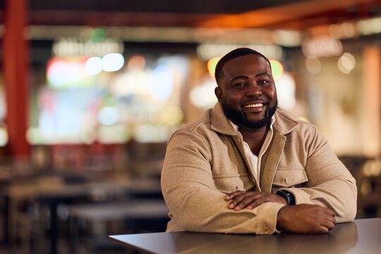 African-American Man Smiles While Sitting At Table In Modern Cafe, Warm Nighttime Ambience, Casual Professional