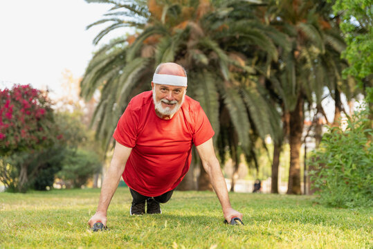 Sportive senior man in red shirt and headband doing push-ups on handles in outdoors park