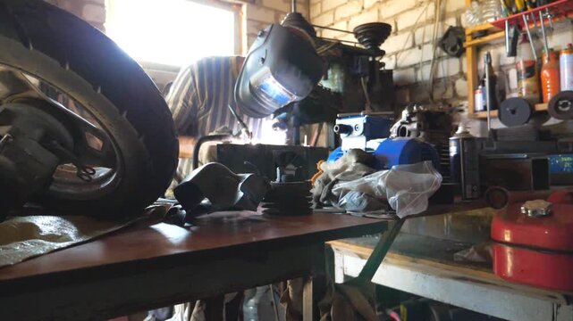 Repairer welding some detail of auto. Mechanic working in garage or workshop with welding machine and other professional equipment. Welder using protective mask from flashes. Slow motion