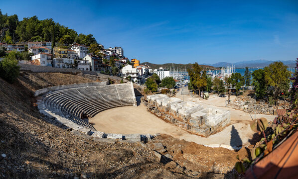 Panoramic view of the ancient Telmessos theatre in Fethiye, Turkey. Scenic cityscape with old stone amphitheater, white houses on a hill and sea port with yachts under a clear blue sky.