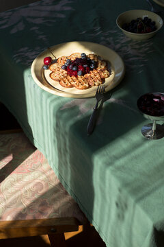 Sunny morning breakfast with round waffle, fresh blueberries, cherries, and strawberry jam on a green tablecloth."
