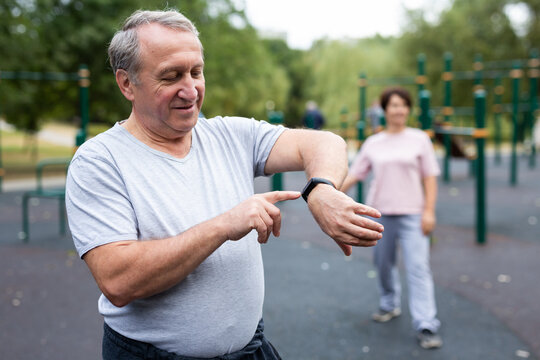 Ssenior man in sportswear looks at fitness watch or at pedometer while exercising in city park