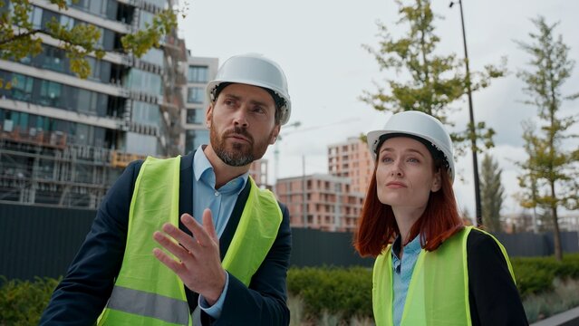 Contractors two engineers man woman talking looking at blueprint building plan diversity people colleagues builders architects wearing safety uniform protective helmets hardhats architecture in city
