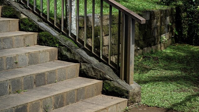 Stone stairs with a metal handrail leading through a lush garden. The scene features a mossy stone retaining wall and green grass, highlighting peaceful outdoor landscape architecture.