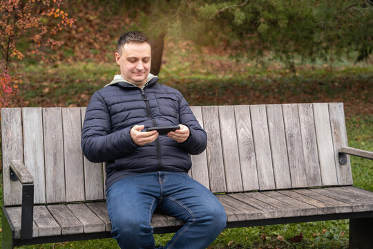 Beautiful 35 year old caucasian man sits on a bench in a city park and looks at his phone. Concept of connection, communication, and technology. Leisure and online communication.