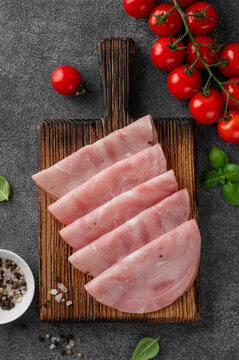 Delicious ham cut into round slices on a cutting board with spices, cherry tomatoes and basil leaves on a dark stone background. Top view.