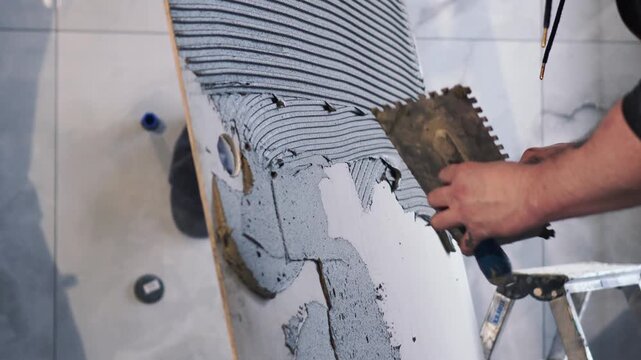 Master tiler spreads glue on huge large format tiles of XXL size. Notched trowel, professional tile work, bathroom renovation, laying blue tiles, slow motion