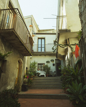 Graniti, Italy - 11 April 2025: View of a narrow, sun-drenched alleyway where aged stone buildings rise above vibrant flora and weathered steps, hinting at timeless Sicilian charm.