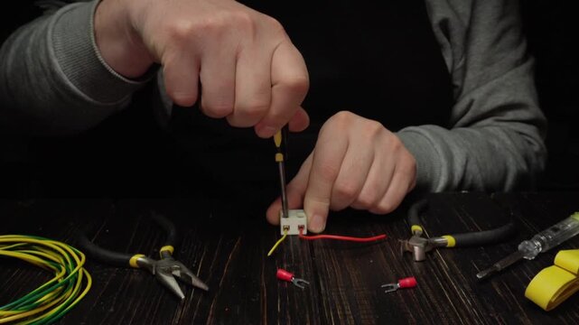 Hands assembling electrical connections with screwdriver, wires, and tools on wooden work surface in a well-lit indoor setting