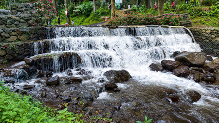 A serene multi-tiered waterfall gracefully cascades over dark, mossy rocks amidst a vibrant green natural park, creating a tranquil and refreshing outdoor escape © Prime