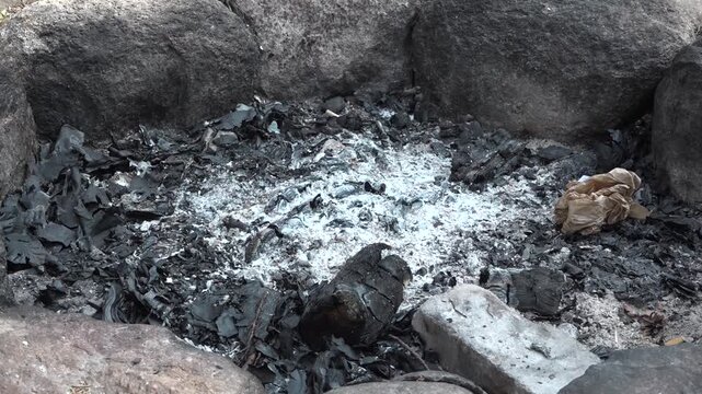 Circular stone fire pit filled with ash and charred remains, surrounded by blackened leaves and debris, showcasing the aftermath of a recent fire in a natural outdoor setting