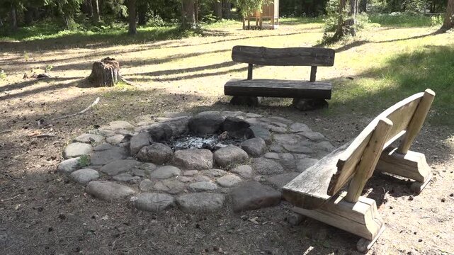 Stone fire pit surrounded by wooden benches in a forest clearing, showcasing the natural setting with sunlight filtering through the trees and shadows on the ground