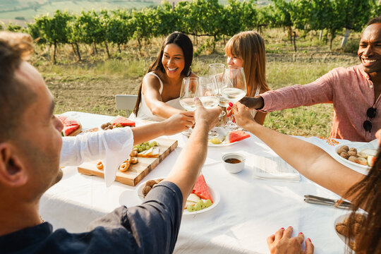 Happy adult friends having fun cheering with white wine together - Multiracial people doing party at summer time in countryside resort with vineyard in background - Main focus on hands
