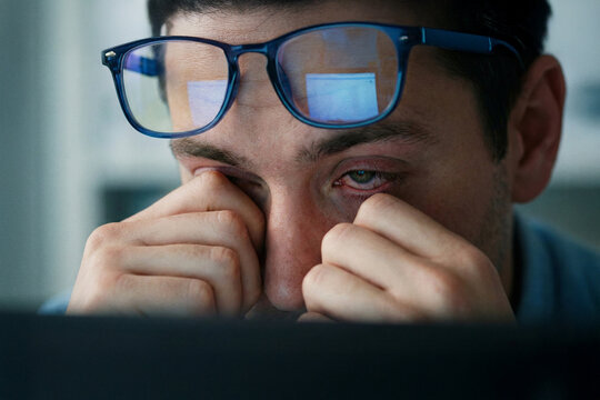 Close-up of an exhausted man with glasses rubbing his tired red eyes, computer screen reflection, digital eye strain and overwork concept