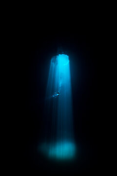 View of a lone diver descends through a shaft of ethereal blue light piercing the inky blackness of an underwater cave, a serene yet dramatic scene, Quintana Roo, Mexico.