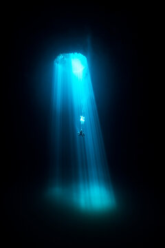View of a diver suspended in the dark abyss as beams of turquoise light pierce through a cenote, illuminating the underwater cave, Tulum, Quintana Roo, Mexico.