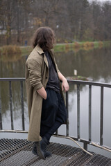 Fototapeta premium Person in a trench coat leaning on a railing by a calm lake in an autumn park, looking away. Moody outdoor portrait with soft background and reflective water.