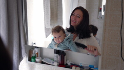 Mother brushing her hair while holding baby, sibling watching in bathroom mirror, family routine,...