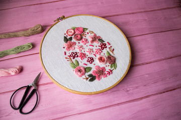 Floral embroidery in wooden hoop with threads, scissors and linen fabric on pink wooden background with copy space. Handmade needlework, DIY craft hobby, sewing and textile art concept.