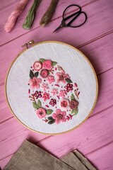 Floral embroidery in wooden hoop with threads, scissors and linen fabric on pink wooden background . Handmade needlework, DIY craft hobby, sewing and textile art concept.