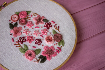 Floral embroidery in wooden hoop with threads, scissors and linen fabric on pink wooden background . Handmade needlework, DIY craft hobby, sewing and textile art concept.