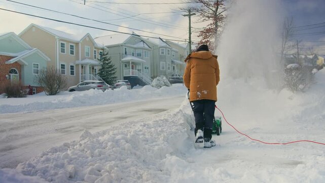Outdoor winter scene: middle-aged homeowner uses powerful wheeled snow blower to clean snow-covered backyard. Snow is thrown to sides, practical snow removal process in residential area.