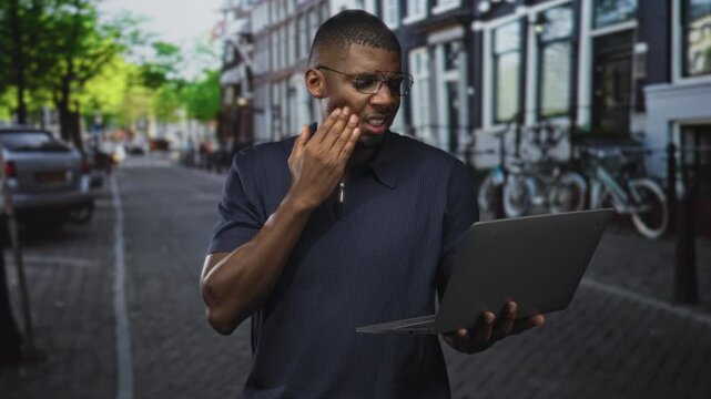 Man holding laptop touching cheek on a cobbled street with parked bicycles and cars; toothache frustration.