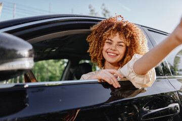 smiling woman with curly hair sitting in black car window, enjoying sunny day outdoors, casual white blouse, cheerful mood, natural light