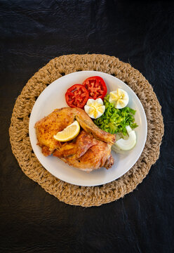 Vista cenital de pollo asado al lim&oacute;n con ensalada de lechuga, tomate, cebolla y huevo. Servido sobre plato blanco y posafuentes de rafia en fondo negro. Ideal para contenido de nutrici&oacute;n.