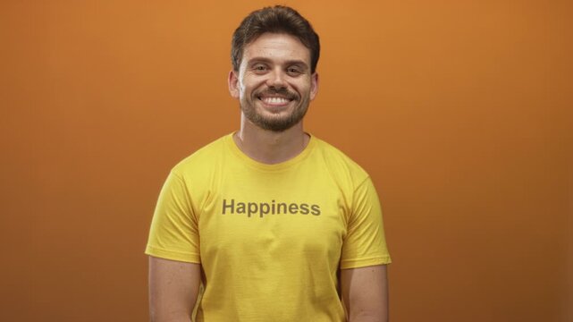Man in yellow t shirt with happiness text sticking out tongue with cheeky expression, seated in studio with orange backdrop; playful joy.
