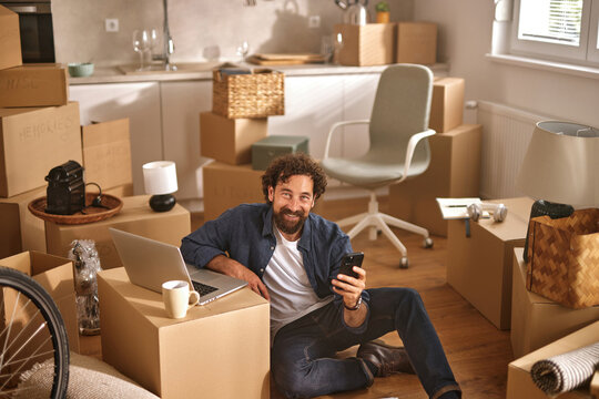 A person sits on the floor surrounded by cardboard boxes in a newly moved-in home while using a laptop and phone, reveling in the thrill of settling in.