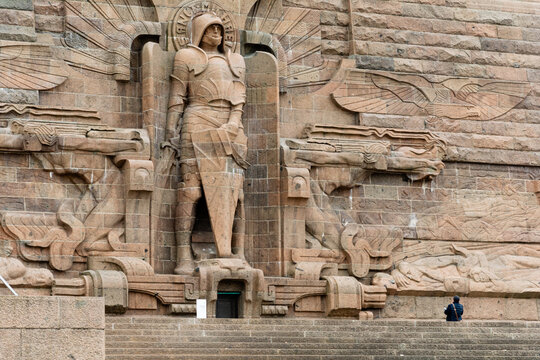 View of a towering stone monument with an armored knight figure stands guard above a lone individual sitting on the steps, Leipzig, Saxony, Germany.