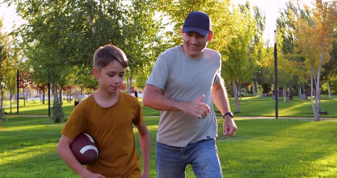 Father runs with son playing American football. Dad coaches American football player. Pair enjoys American football training in park. Healthy lifestyle promotes fitness. Happy bonding time outdoors.