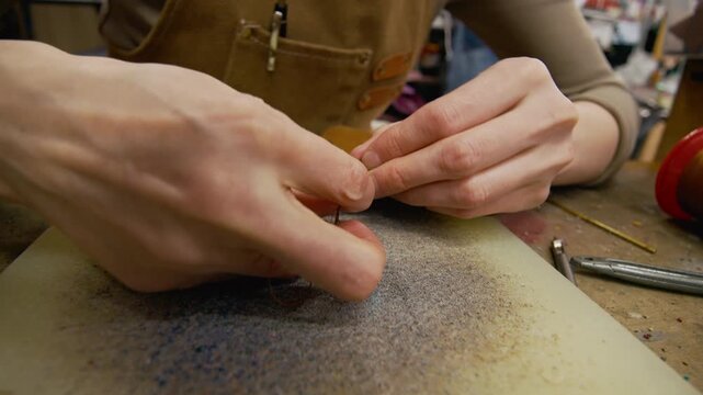 Close up on hands of young craftswoman stitching brown suede panels with waxed thread while sewing leather pieces together at craft table in workshop