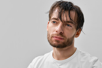 Obraz premium Front portrait of adult short bearded man with wet hair and strands on forehead wearing white t shirt, looking far, light gray minimal studio background.