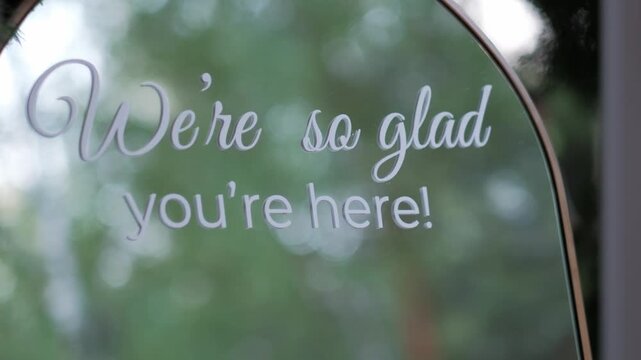 A white cursive sign on a mirror says We're so glad you're here with a blurred green forest background. Static shot.