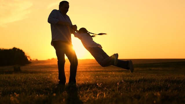 Cheerful people in park, parents children. Happy father daughter twirling in sunny meadow. Family, dad daughter playing, sun park lawn. Father twirling daughter, holding hands, kid girl laughing