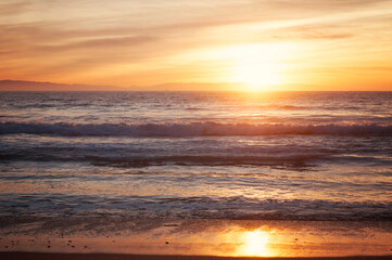 Obraz premium Golden sunset over ocean waves with warm orange sky reflecting on wet sand, creating a peaceful coastal landscape scene with soft light and tranquil atmosphere.