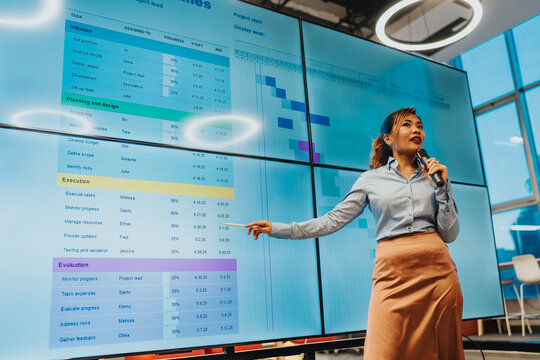 A female presenter speaks into a microphone beside a wall of blue data charts.