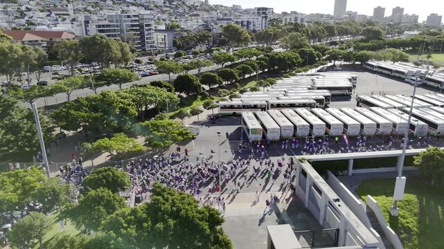 Drone does a 360 around parade proceeding toward stadium, walking past bus depot during Cape Town Minstrels Carnival on sunny afternoon in Green Point, South Africa