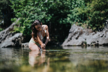 Fototapeta premium A woman in a bikini wades in a shallow river, her hands touching the water. The scene shows a peaceful summer moment by rocks and lush green trees.