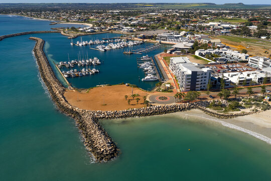 An elevated aerial view of Geraldton marina shows boats moored within a protected harbour, surrounded by coastal buildings, breakwaters, and turquoise water.