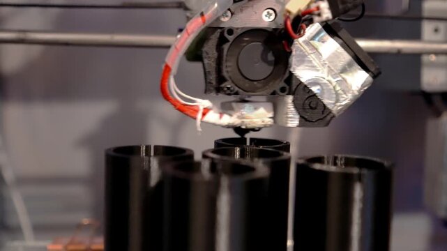 A 3D printer in operation, printing plastic parts using fused filament manufacturing technology. The print head deposits molten material onto a glass work plate, forming detailed black components.