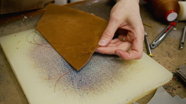 Close up on hands of young female artisan unpicking stitches on brown leather piece using sharp scissors while crafting custom purse in workshop