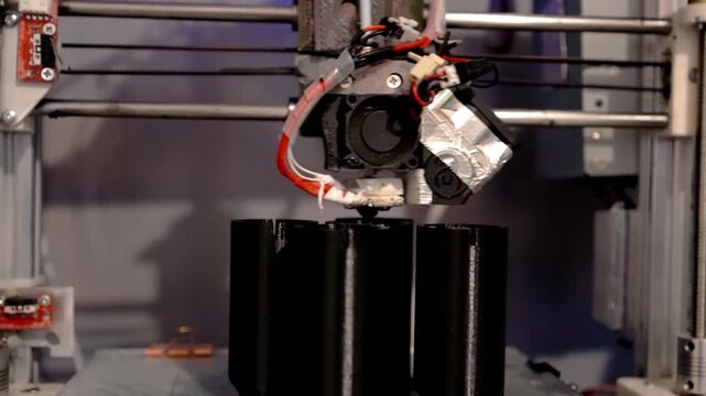 A 3D printer in operation, printing plastic parts using fused filament manufacturing technology. The print head deposits molten material onto a glass work plate, forming detailed black components.