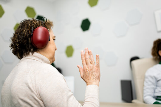 A 60-year-old woman undergoes hearing testing at a medical center. The woman is having an audiometry test with headphones. Photo taken from behind as the woman raises her hand.